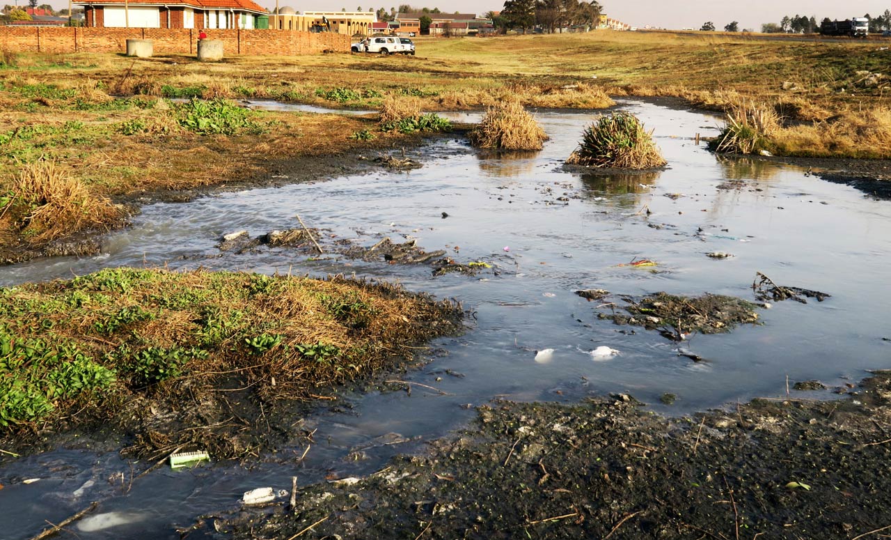 A stream of untreated sewage flowing from a broken pump station in Evander, Mpumalanga. The white vehicles in the background belong to a municipal crew who were restoring electric cables to the pump station, which had been stolen.